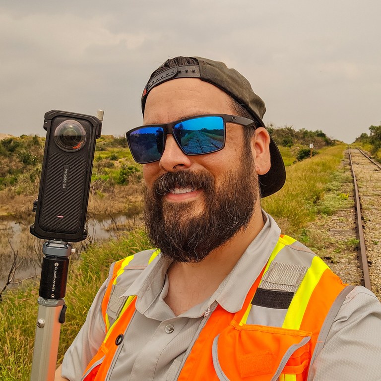 Robert Robinson wearing field safety gear beside a rail line, holding a 360° camera